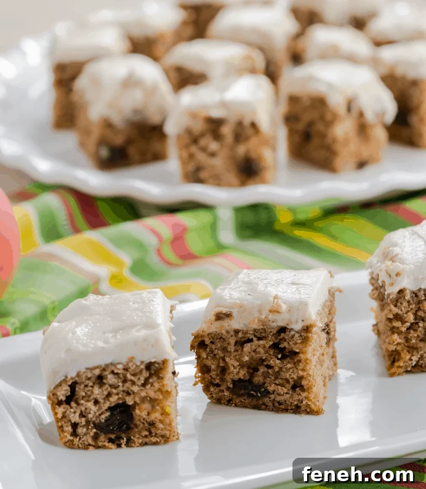 Three raisin spice cake bars frosted with brown sugar cream cheese frosting. Cinnamon sticks on white plate.