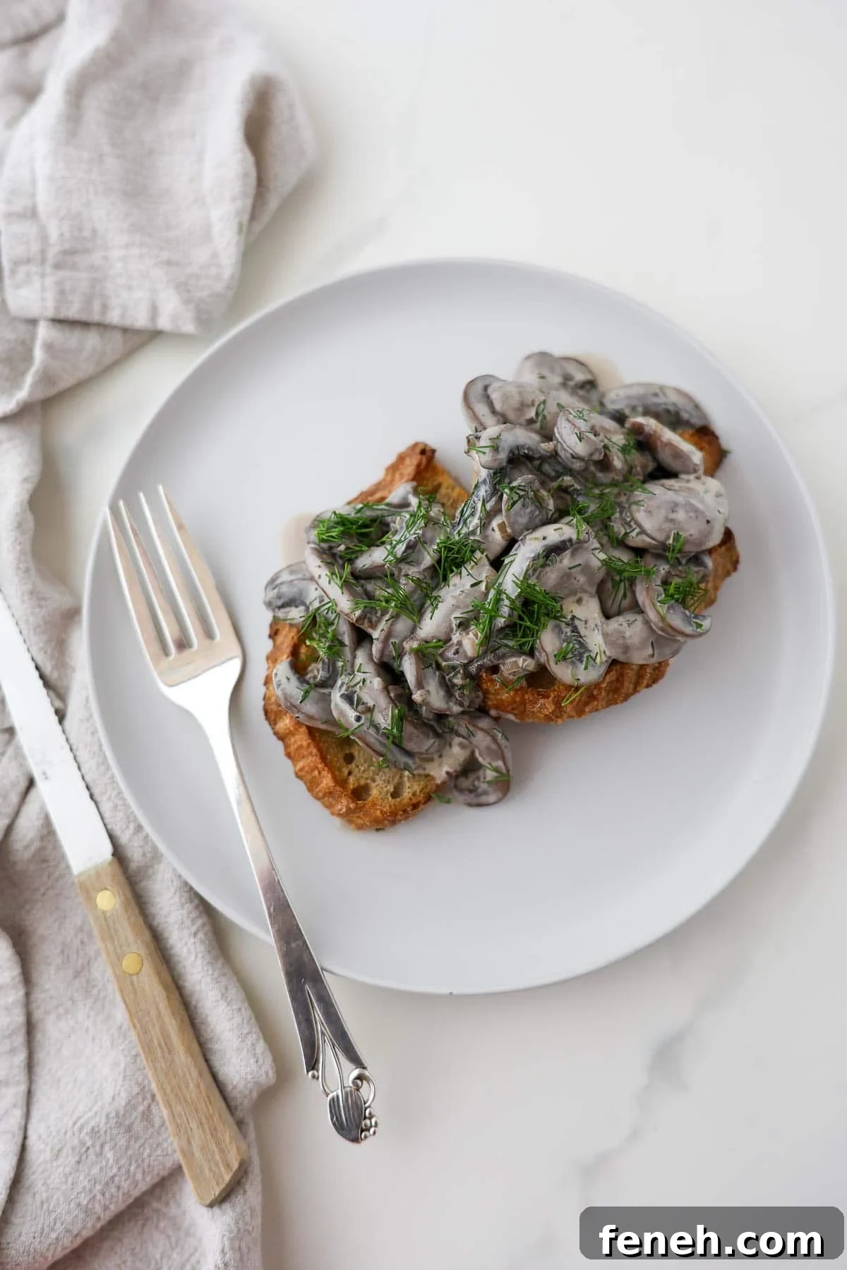 Scandinavian mushroom toast on a white plate next to a fork and knife.