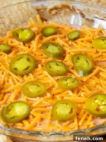 A glass pie dish filled with cream cheese and salsa dip, topped with shredded cheddar cheese and sliced jalapeños, surrounded by tortilla chips.