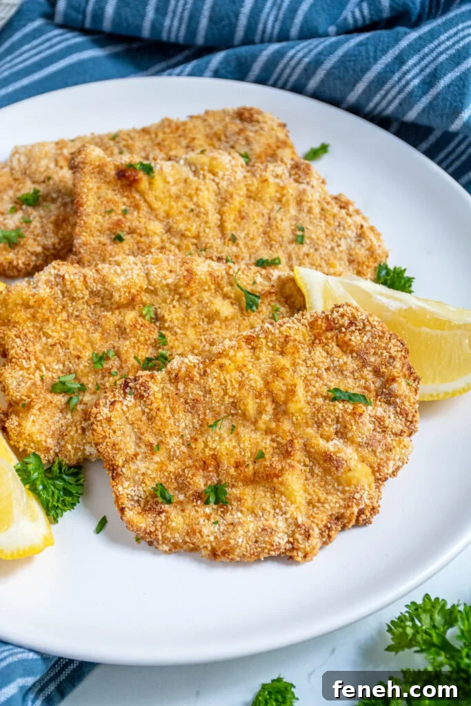 Close up of a row of air fryer pork schnitzels on a white plate with parsley and lemon wedges.