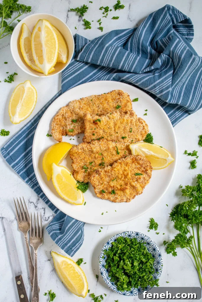 Overhead view of a tabletop with a white plate full of air fryer pork schnitzel garnished with fresh parsley and surrounded by lemon wedges.