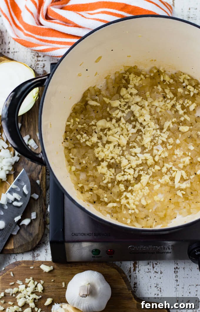 A black Dutch oven holds sauteed onions and chopped garlic. An orange and white striped towel, a chopped raw onion and chopped raw garlic sit next to the pan.