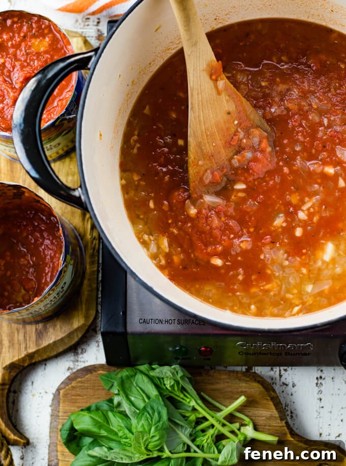 A black Dutch oven with sauteed onions and garlic, vodka sauce and crushed tomatoes. Cans of tomatoes and fresh basil sit next to the pan.