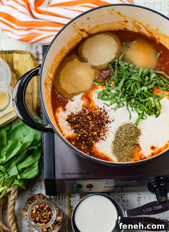 A black Dutch oven has a tomato base with spices, herbs and whole cream on it. Cream, fresh basil and orange and white striped napkin sit next to the pan.