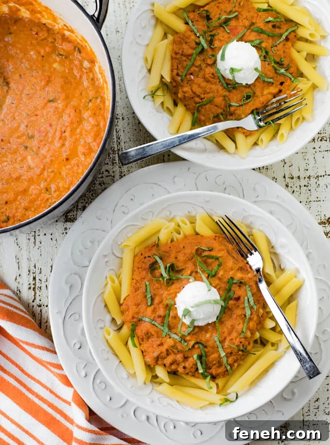 Vodka Pasta Sauce sits on top of penne pasta with a dollop of ricotta cheese and julienned basil leaves are scattered on top. A pan of the sauce and another bowl is blurred in the background.