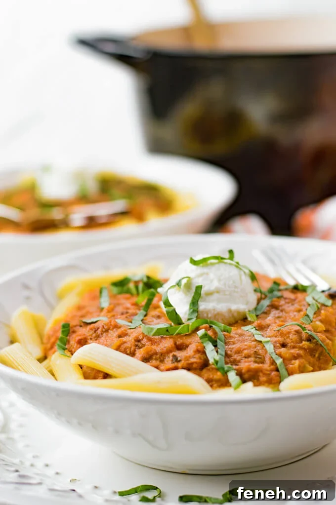 Vodka Pasta Sauce sits on top of penne pasta with a dollop of ricotta cheese and julienned basil leaves are scattered on top. A pan of the sauce and another bowl is blurred in the background.