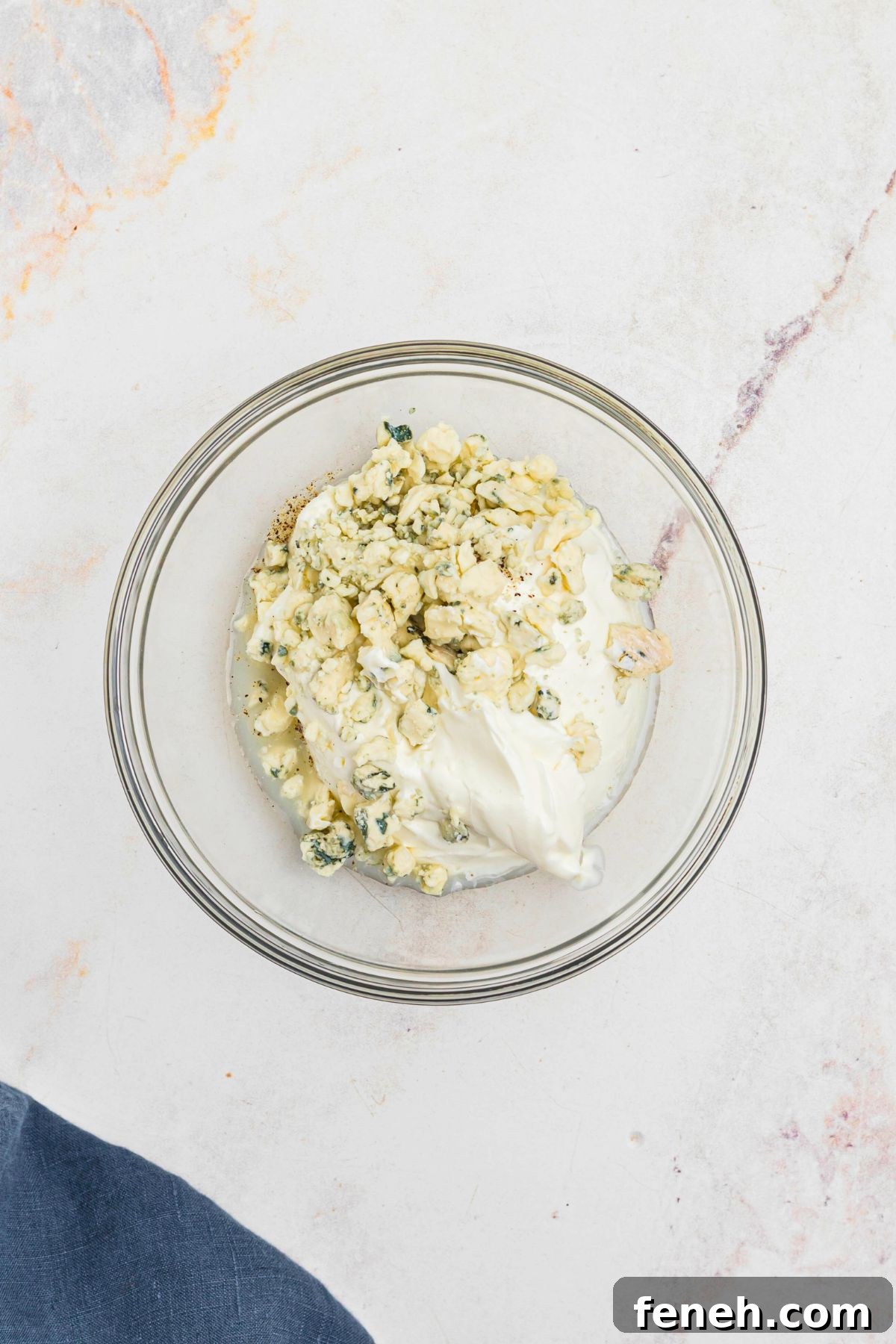 Clear glass bowl with sour cream, blue cheese, and other ingredients, before being mixed together. 