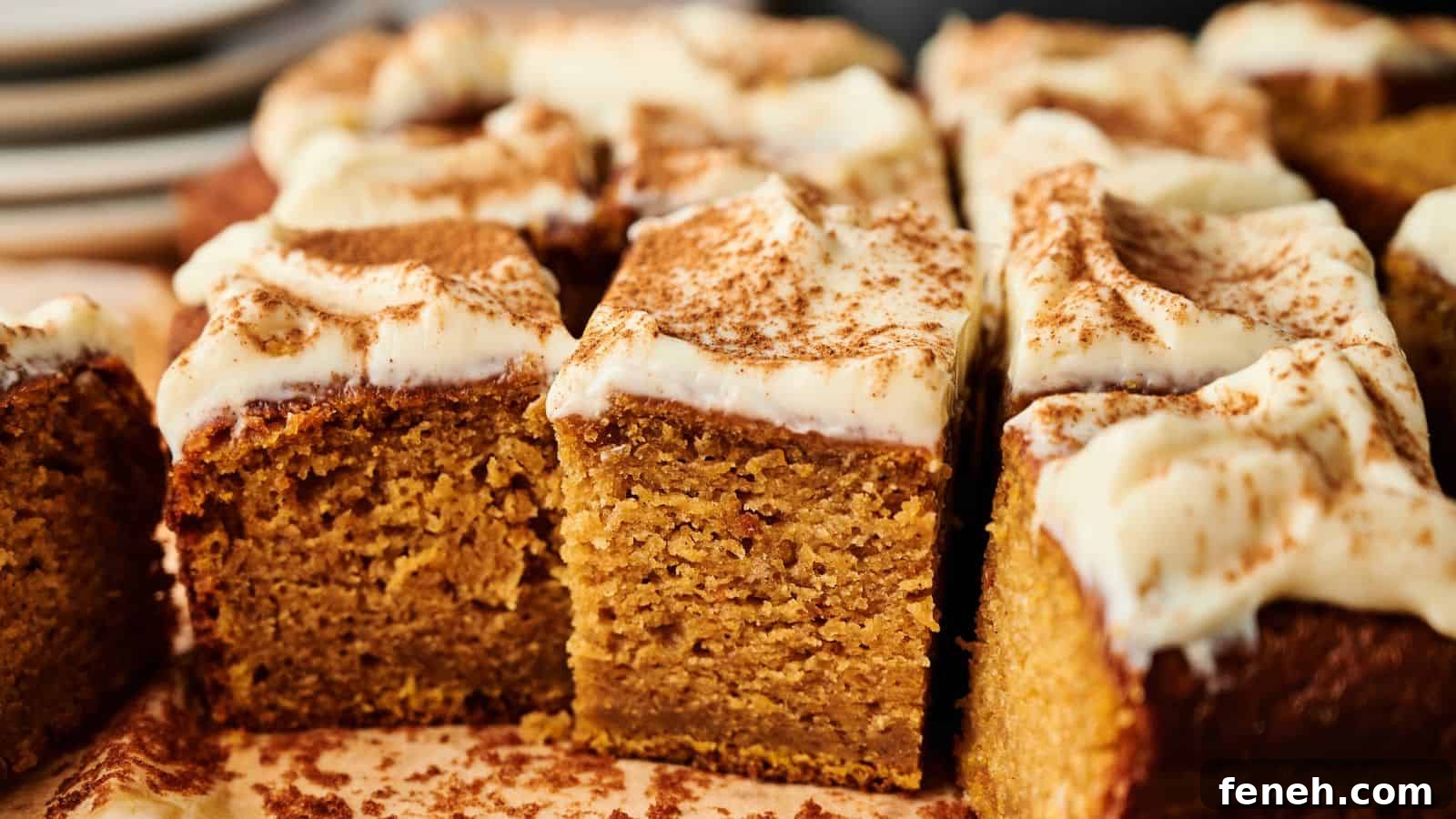 Close-up of sliced pumpkin cake topped with cream cheese frosting and a dusting of cinnamon, with plates in the background.