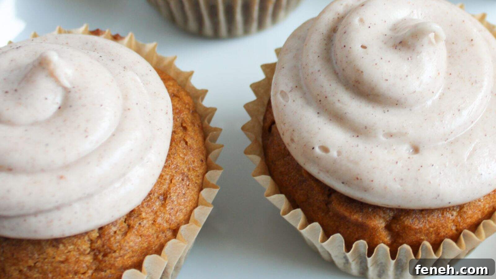 Two cupcakes with light brown frosting are shown on a white surface, each in a beige paper liner.