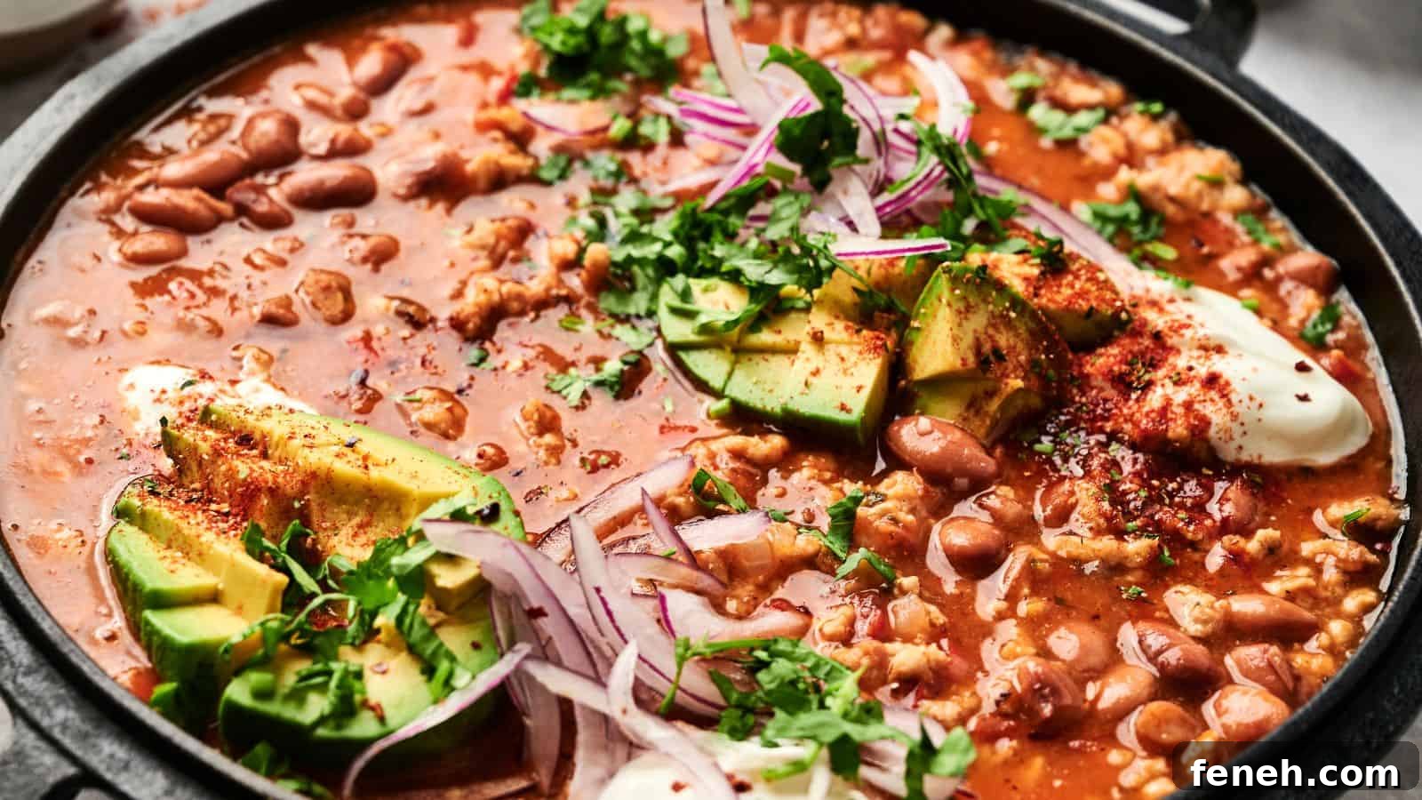 A close-up of a pot of chili with beans, avocado slices, red onion, sour cream, and garnished with chopped cilantro.