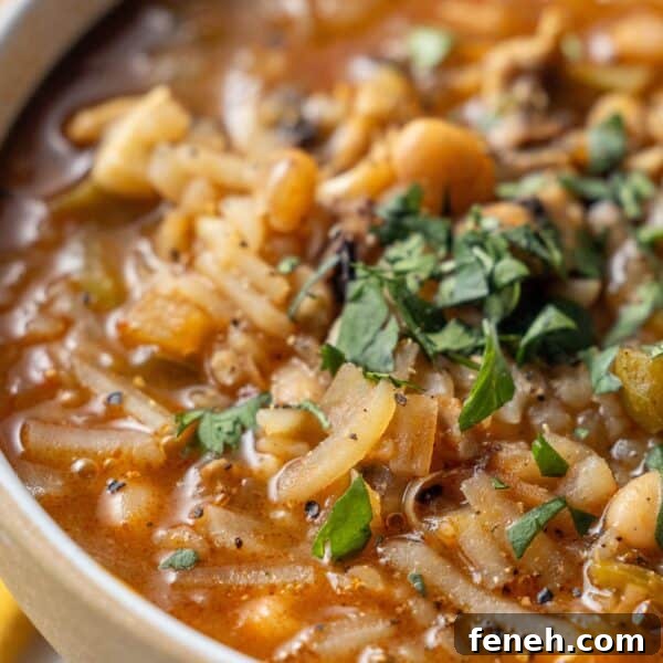 Close-up of a bowl of vegan soup containing noodles, vegetables, beans, and herbs, topped with chopped fresh parsley and black pepper.