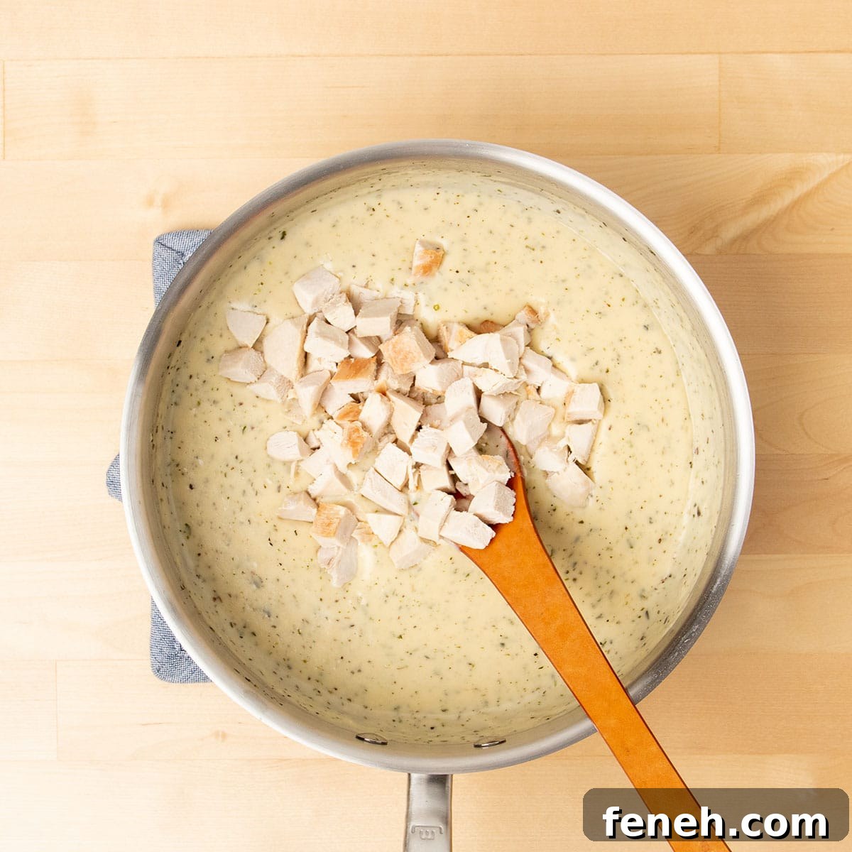 light brown cream soup with herb flecks with brown mixing spoon and cubed cooked chicken in same saucepan