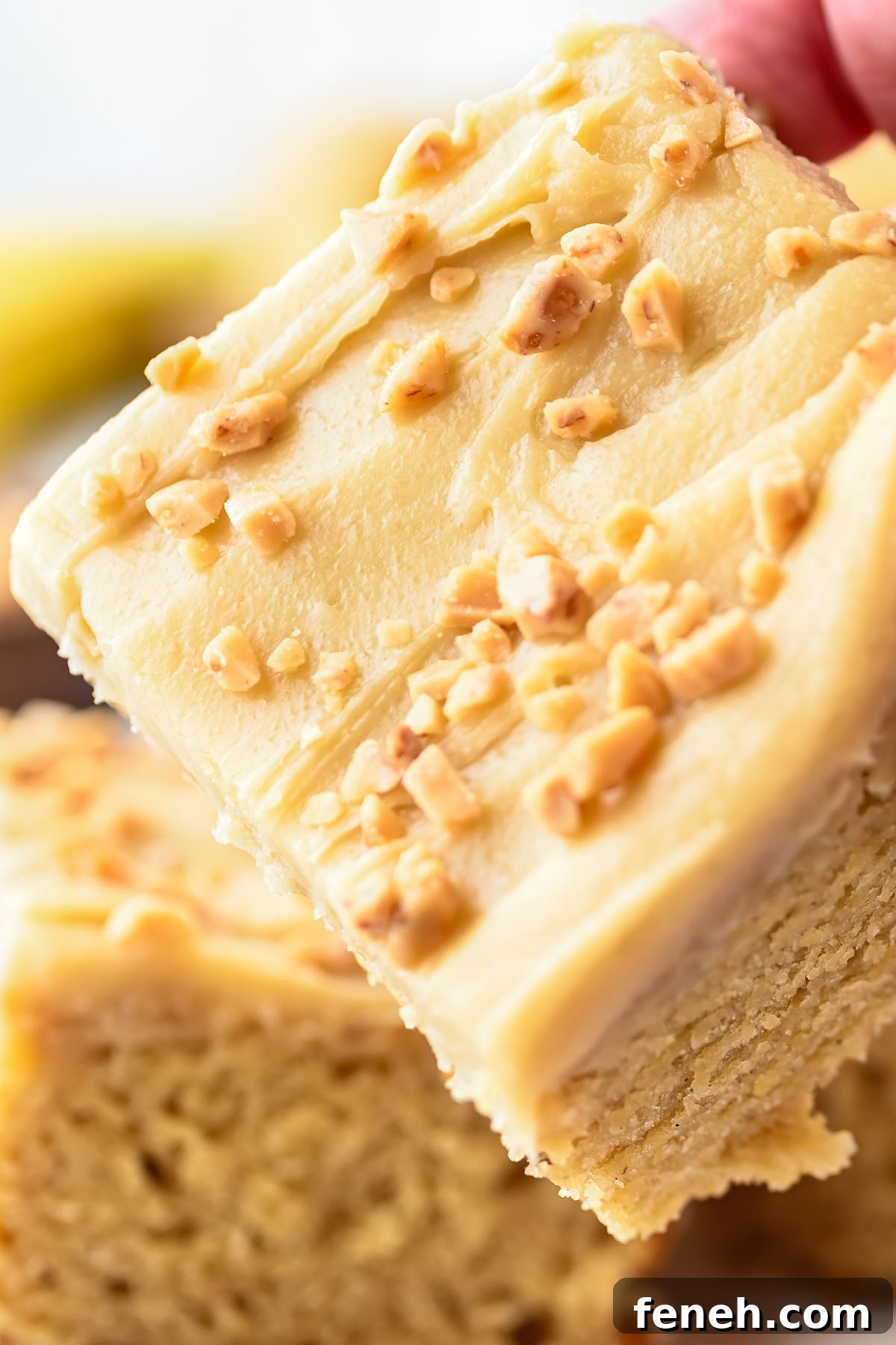 A hand holding a slice of banana blondies with brown butter frosting topped with chopped nuts.