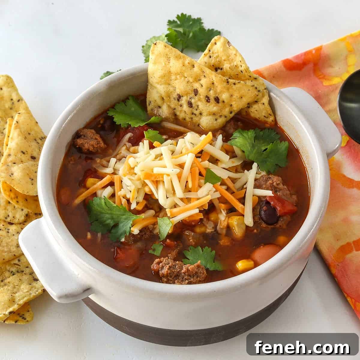 Beef chili soup in a white bowl with shredded cheese garnishes.
