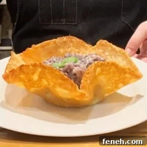 A person presenting a dish with crispy edges shaped like a bowl on a white plate. The bowl contains a filling with a basil garnish. They are wearing a dark apron, and kitchen items are visible in the background.