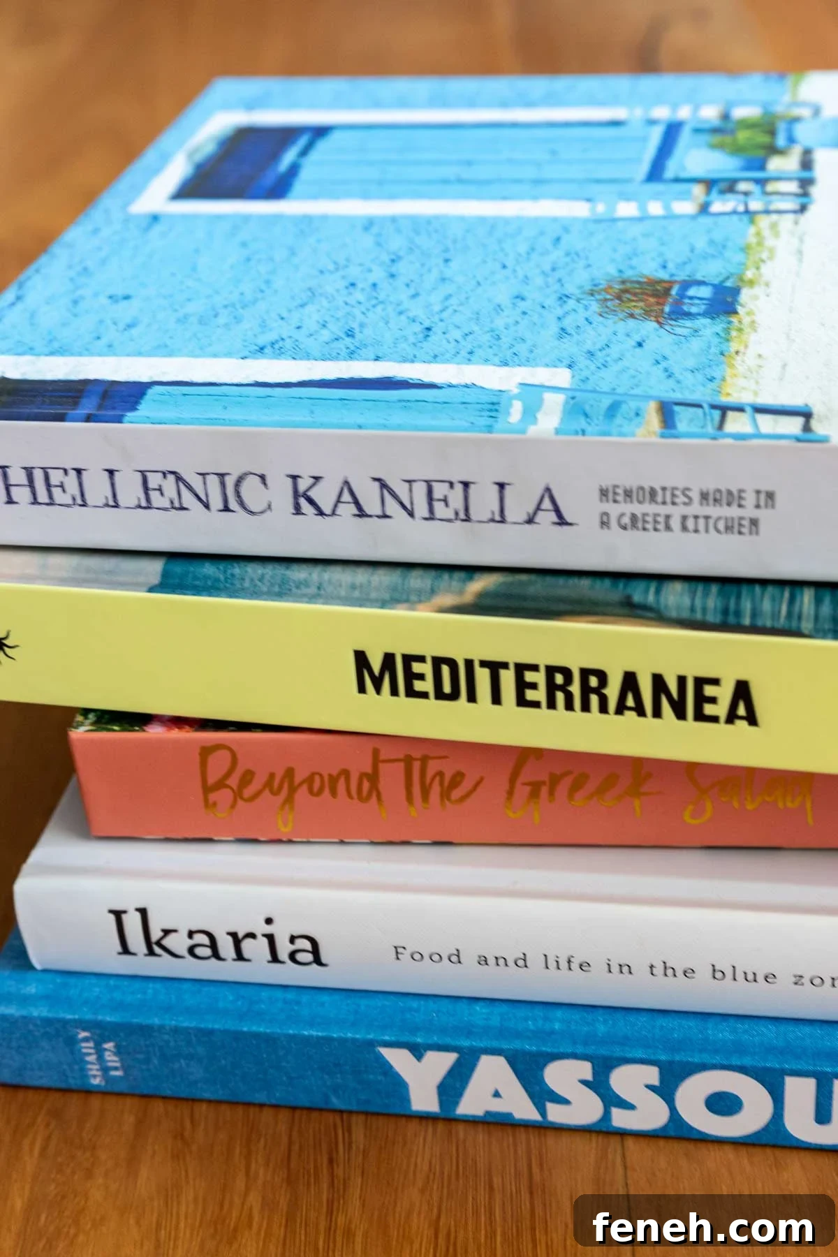 A small stack of Greek cookbooks on their sides, on a wooden table.