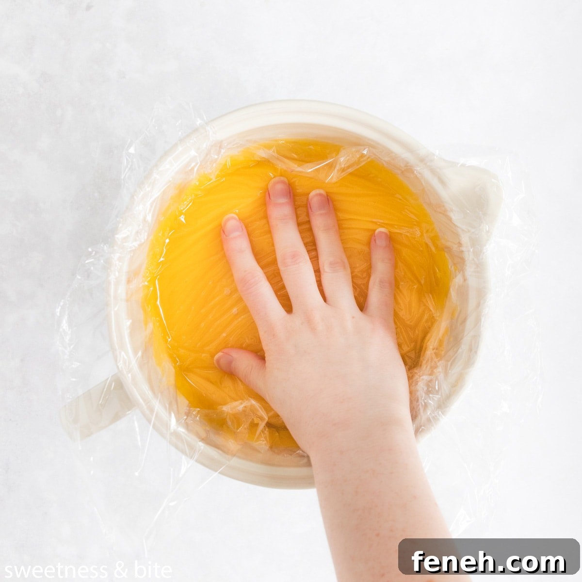 Lemon curd in a large jug, with a hand pressing plastic wrap onto the surface.