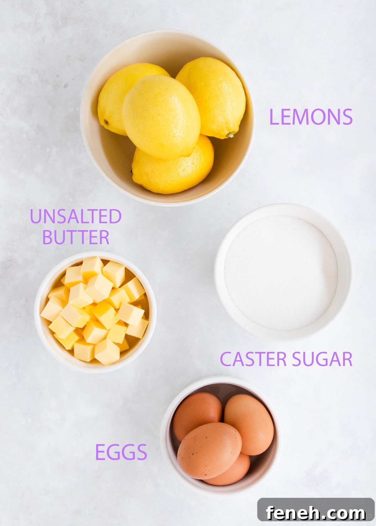The lemon curd ingredients in white bowls on a grey background - lemons, butter, sugar and eggs.
