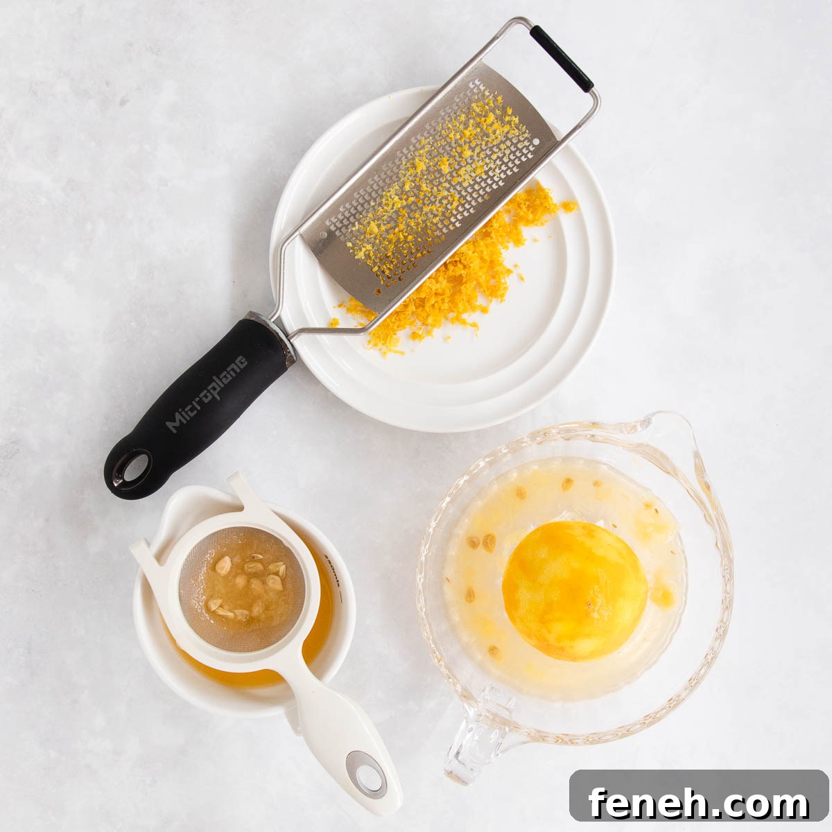 Lemon zest on a white plate with a microplane grater, and a glass lemon juicer with a juiced lemon, on a grey marble background.