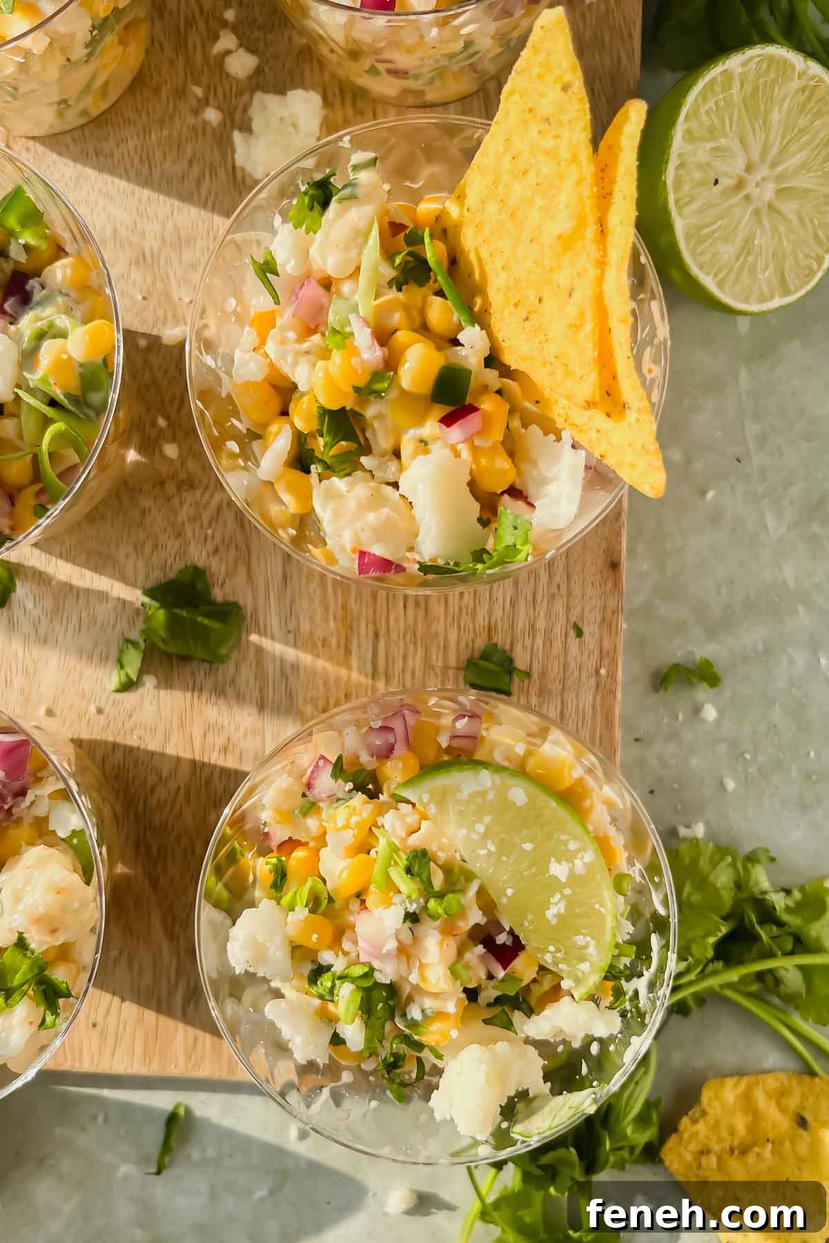 elote in small plastic cups garnished with lime wedges and tortilla chips on wood cutting board.