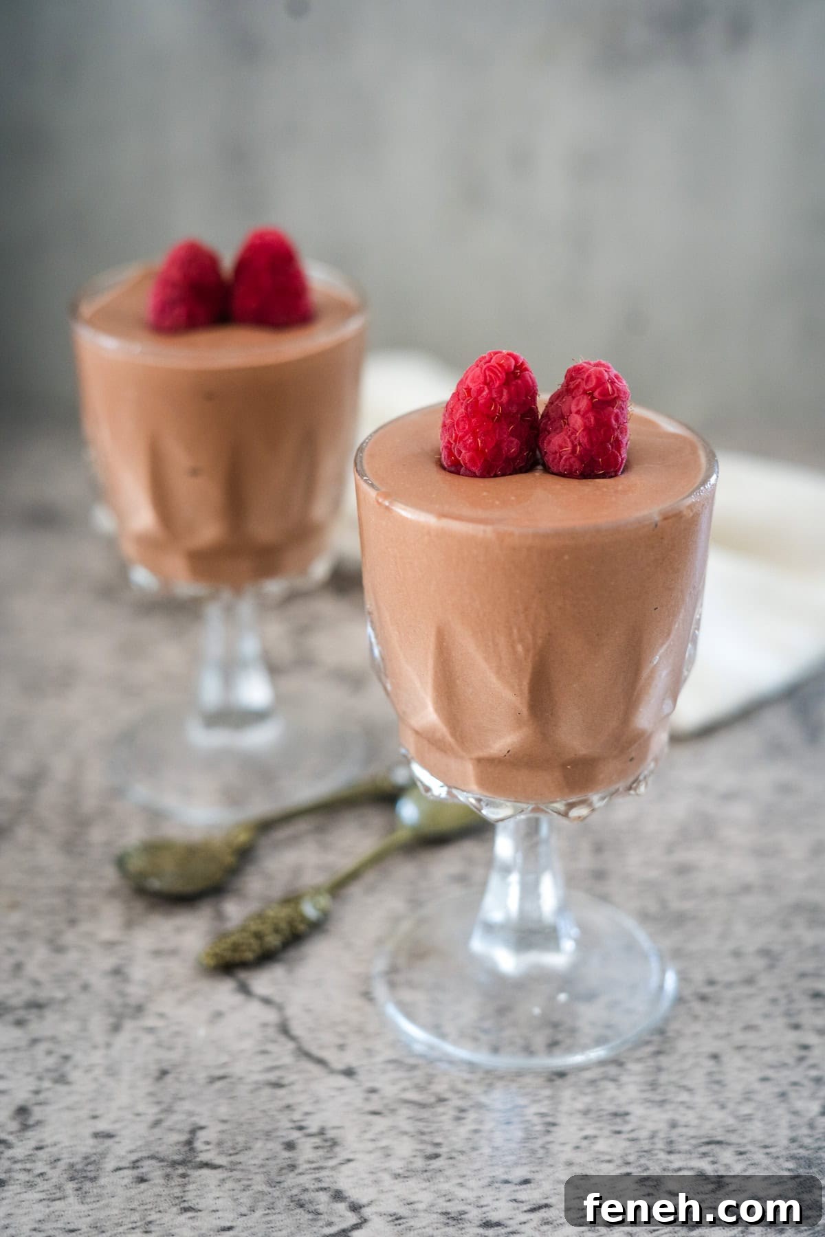 Two glass cups of chocolate mousse topped with raspberries, placed on a textured surface with two small spoons between them.