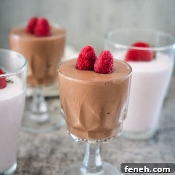 Four dessert glasses with mousse, two with chocolate mousse topped with raspberries and two with pink mousse also topped with raspberries. They are placed on a gray textured surface.
