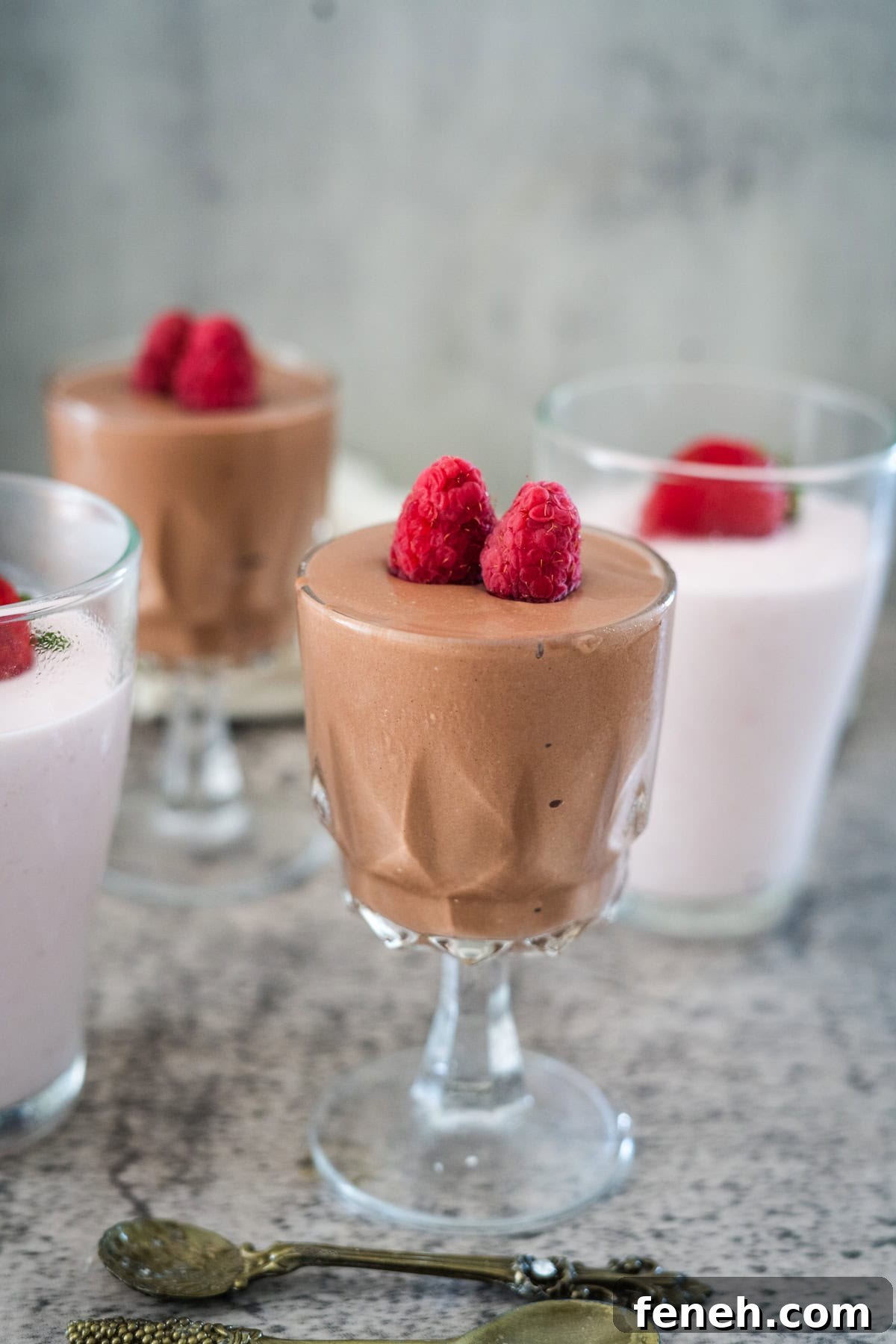 Four glass cups filled with chocolate and strawberry mousse topped with raspberries, arranged on a grey surface with two small spoons nearby.