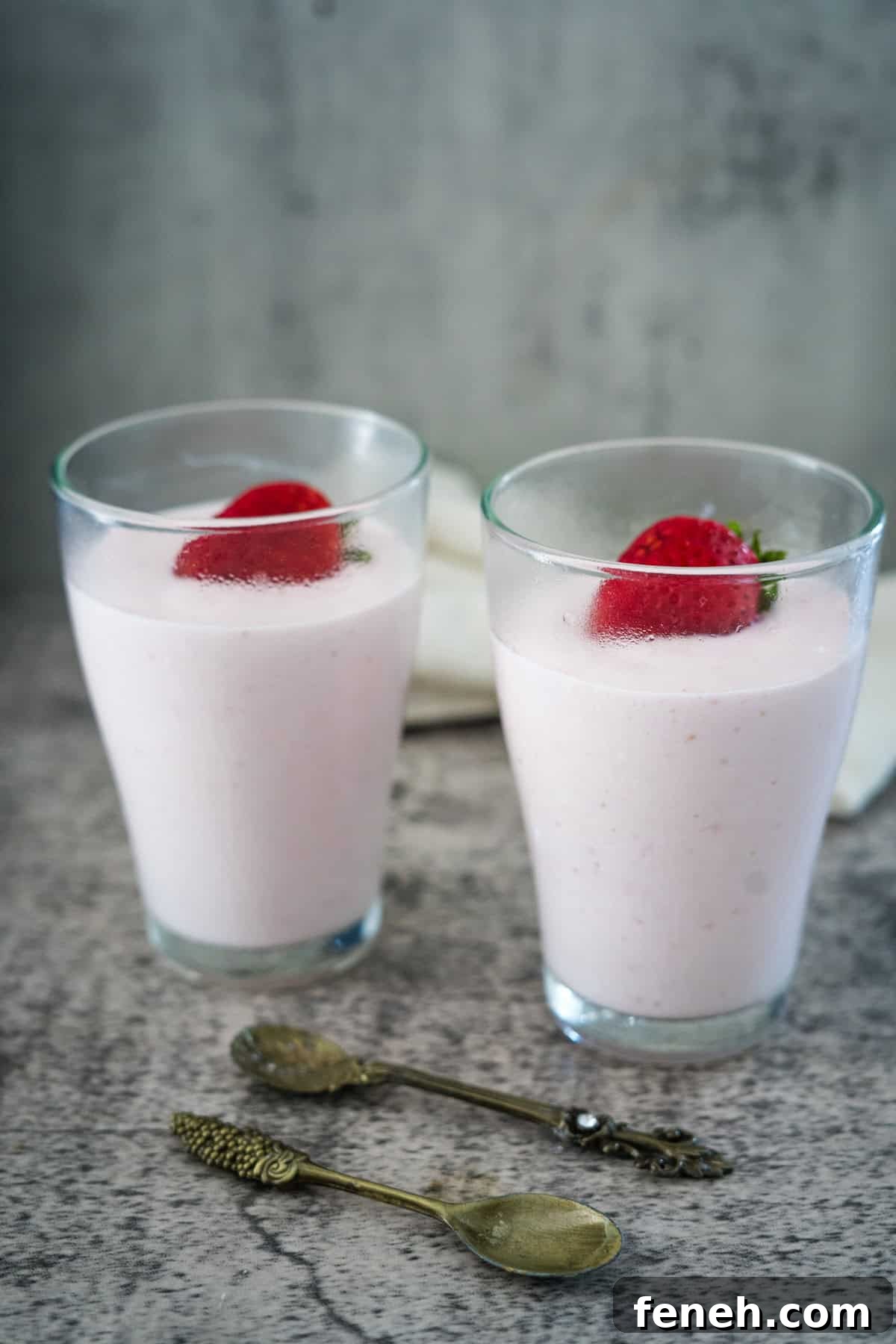 Two glasses filled with pink strawberry smoothies, each topped with a whole strawberry, are placed on a textured surface with two vintage-style spoons in front.