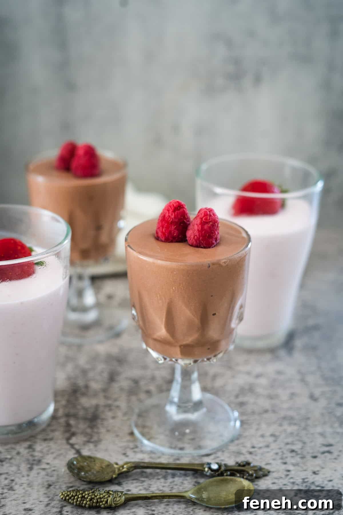 Three glasses of mousse, two chocolate and one strawberry, each topped with raspberries. Two decorative spoons are in the foreground.