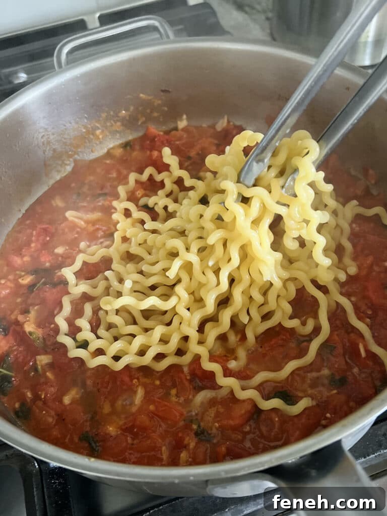 Cooked fusilli being added to pan with tomato sauce.