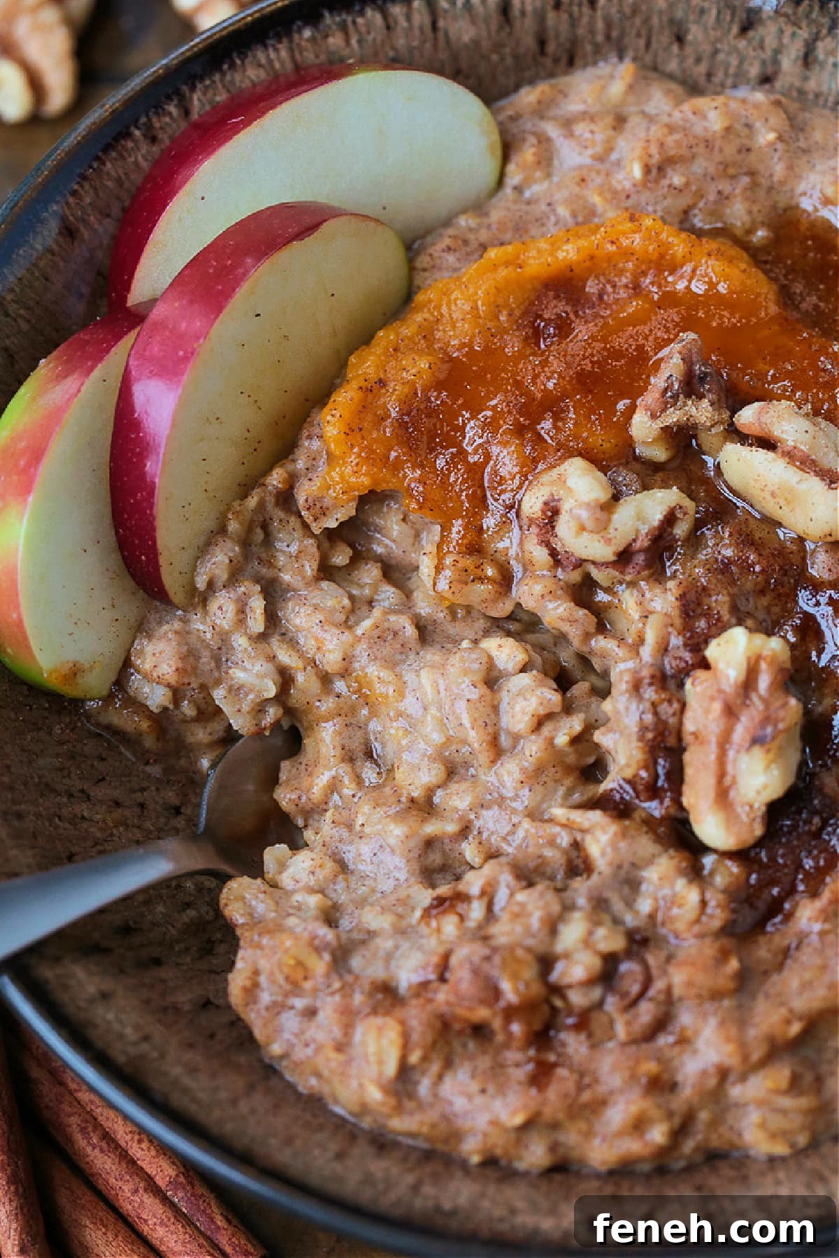 A bowlful of pumpkin oatmeal topped with apples, walnuts and pumpkin puree, with a spoon.
