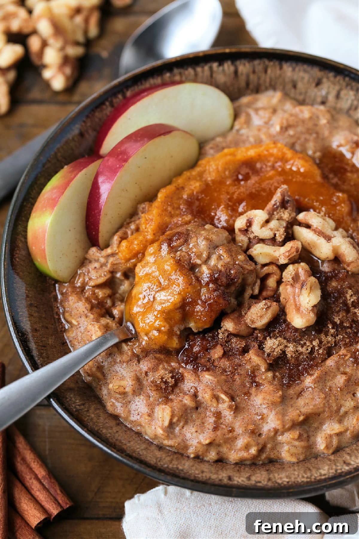 A bowlful of pumpkin oatmeal topped with apples, walnuts and pumpkin puree, with a spoon.