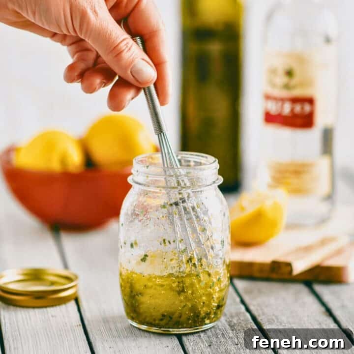 Greek salad dressing with ouzo being mixed in a glass jar with a whisk and fresh herbs.