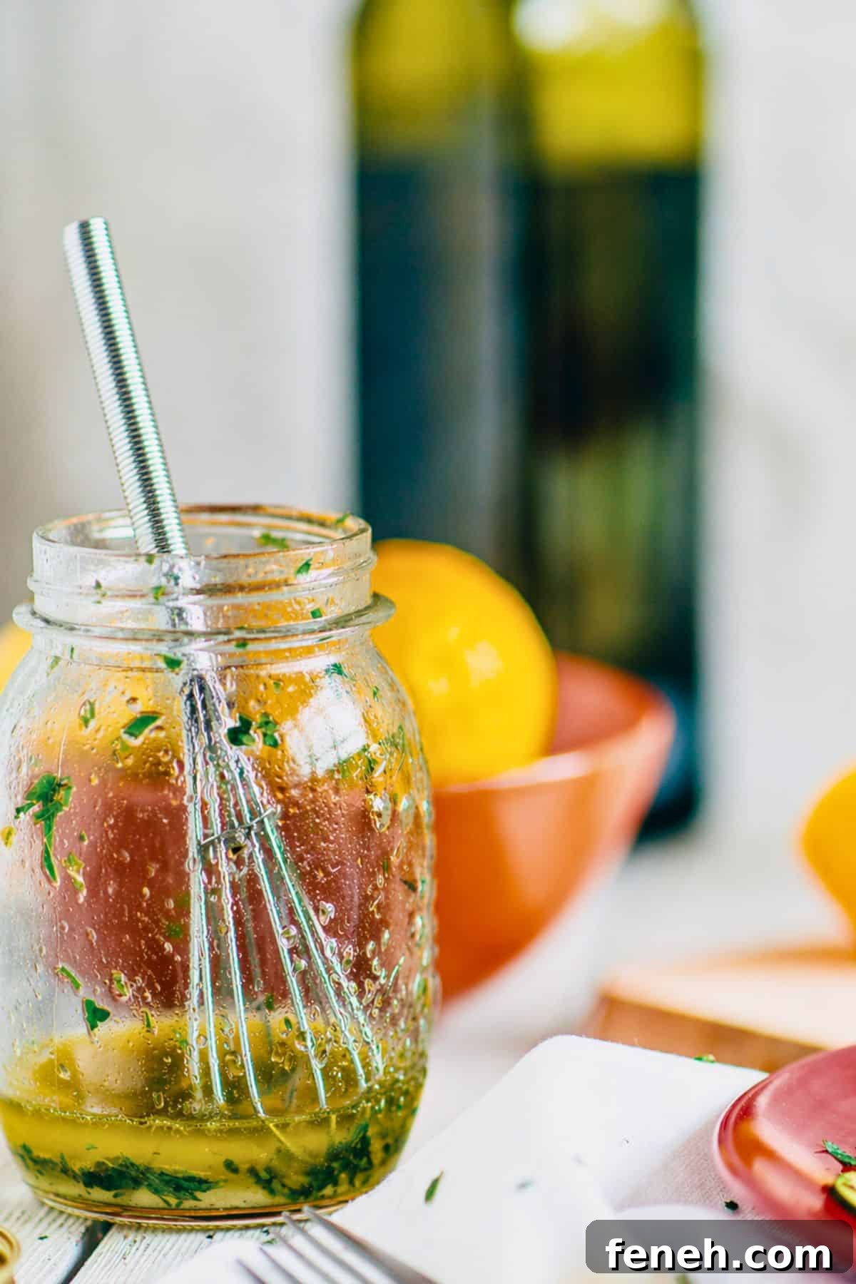 Close up of Greek salad dressing with ouzo in a glass jar with herbs, lemon and olive oil in the background.
