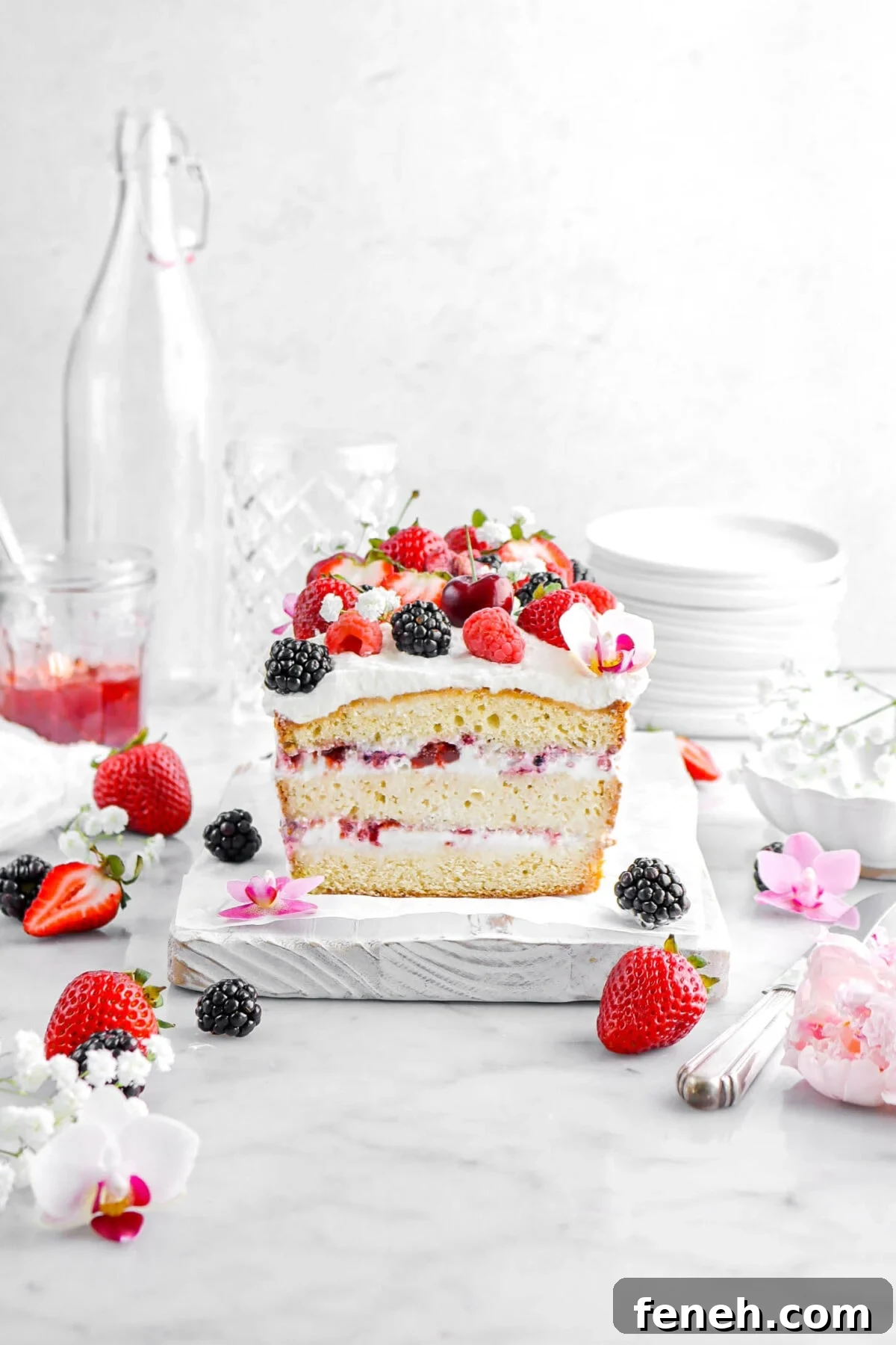 ice cream pound cake on white wood board with berries and flowers around, a jar of jam, empty glasses, and a stack of plates behind.