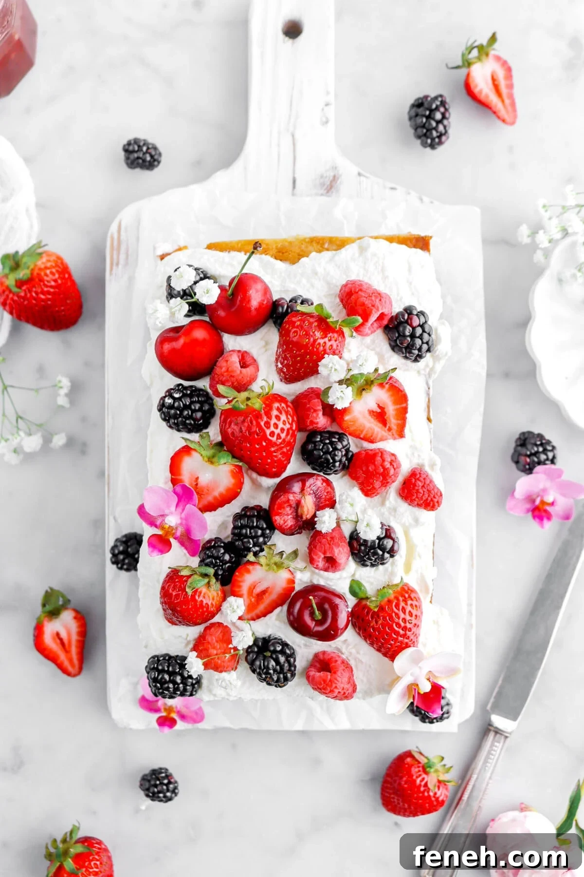 overhead shot of ice cream cake on white wood board with whipped cream, berries, and flowers on top.
