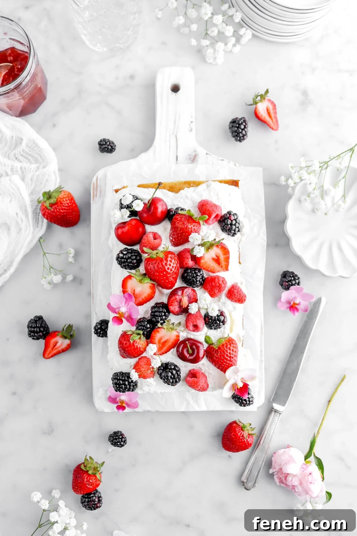 overhead shot of loaf cake on white wood board with whipped cream, fresh berries, and flowers on top with more berries and flowers around, a scalloped bowl, and jar of jam around.