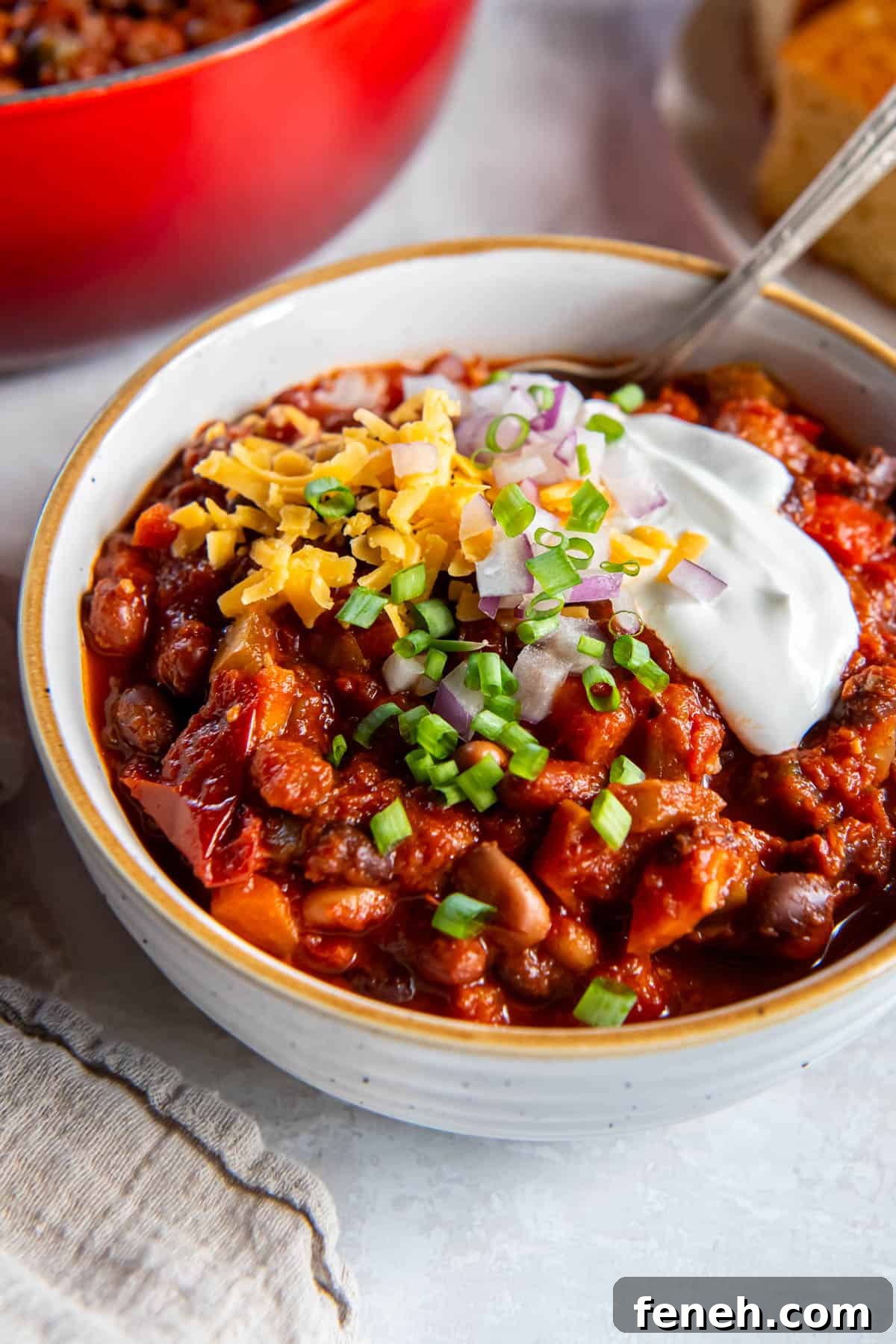 A bowl filled with three bean chili and topped with sour cream, cheddar cheese, red onion and chopped chives. A spoon also sits in the bowl for serving.