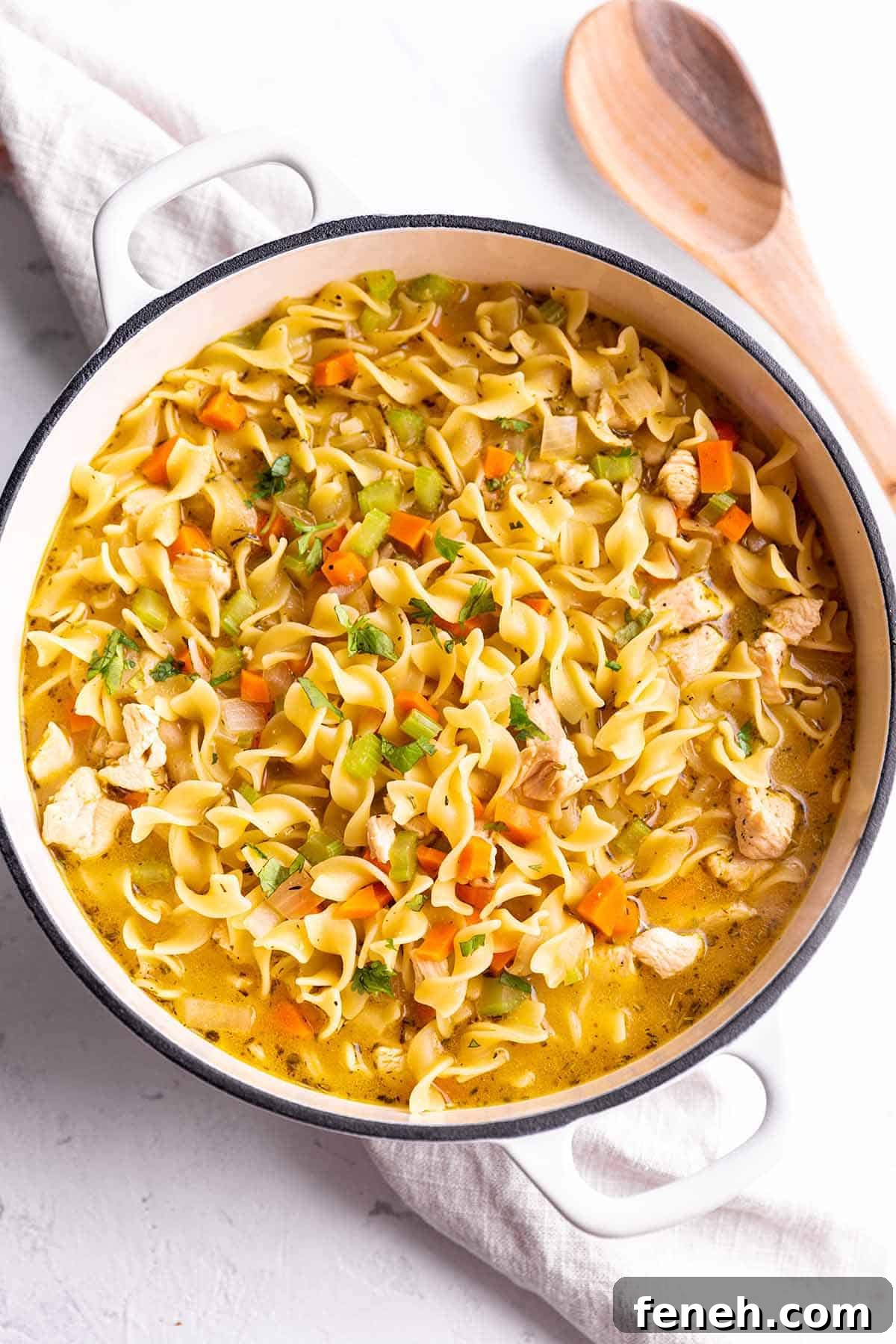 A white Dutch Oven full of a chicken noodle soup sitting on a napkin with a wooden spoon.