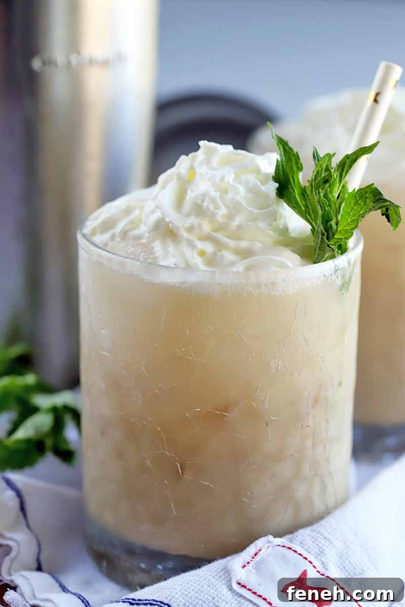 Closeup image of a glass filled with a milky coffee beverage, topped with whipped cream and a sprig of mint, with a white and gold foil paper straw, on a gathered white cloth with more fresh herbs and a stainless steel cocktail shaker with matching lid in the background.