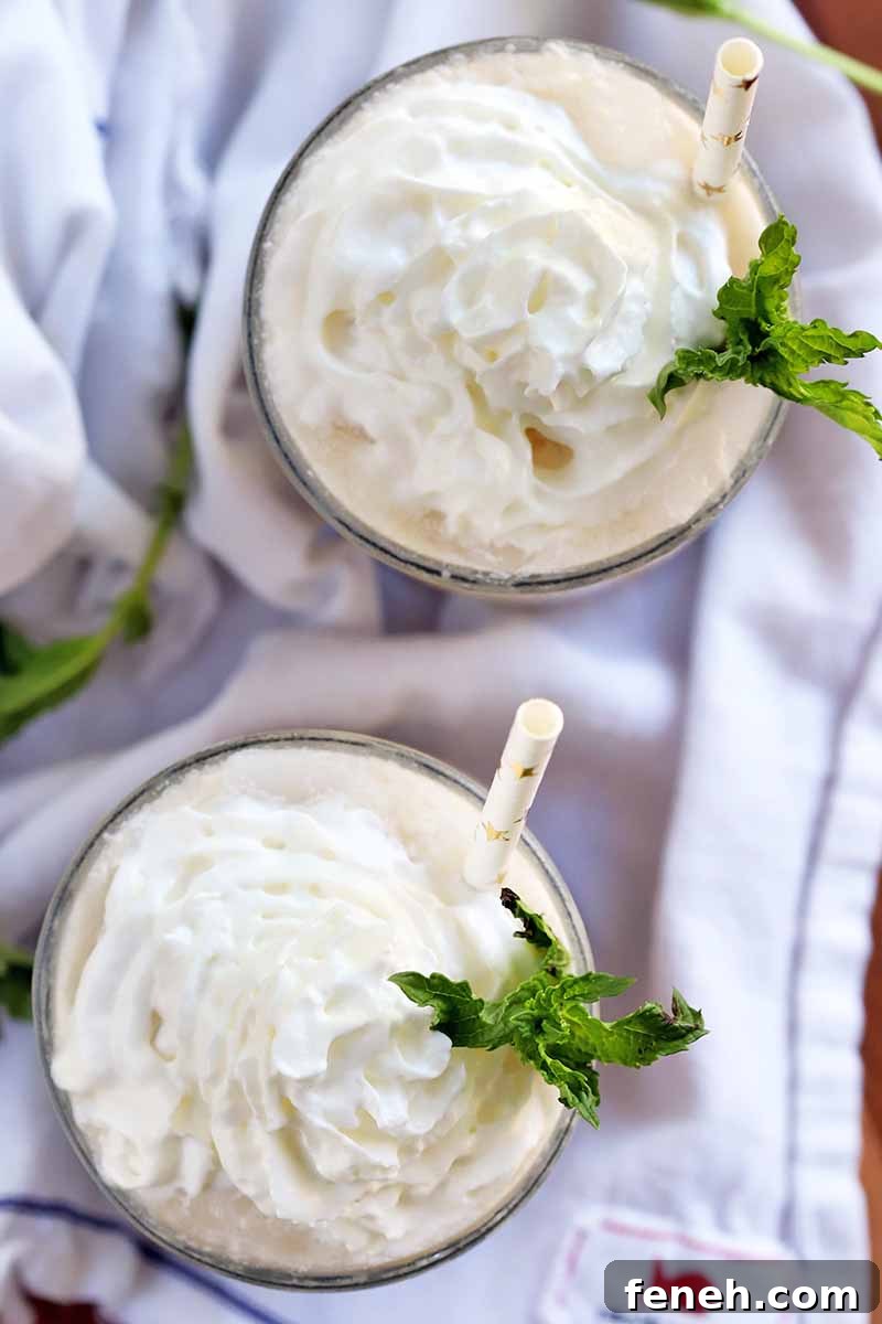 Overhead vertical shot of two glasses topped with whipped cream and sprigs of mint, with white paper straws decorate with gold foil straws, o a folded and gathered white cloth, on a brown wood surface.