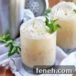 Two lowball glasses filled with a coffee cocktail topped with whipped cream, a paper straw with gold foil decoration, and a sprig of fresh green mint, with more mint, a metal cocktail shaker, and a gathered white cloth, on a brown wood background.