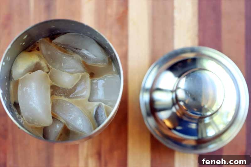 Overhead shot of a stainless steel cocktail shaker filled with ice and coffee, with its lid to the right, on a striped wood surface.
