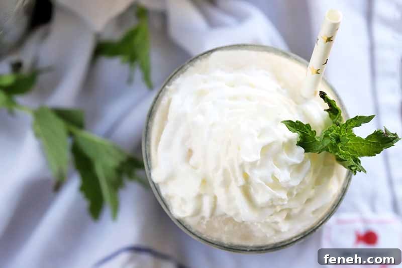 Overhead shot of a glass topped with whipped cream and a sprig of mint with a white straw with gold foil decorative accents, on a gray wrinkled and gathered cloth with more fresh herbs.