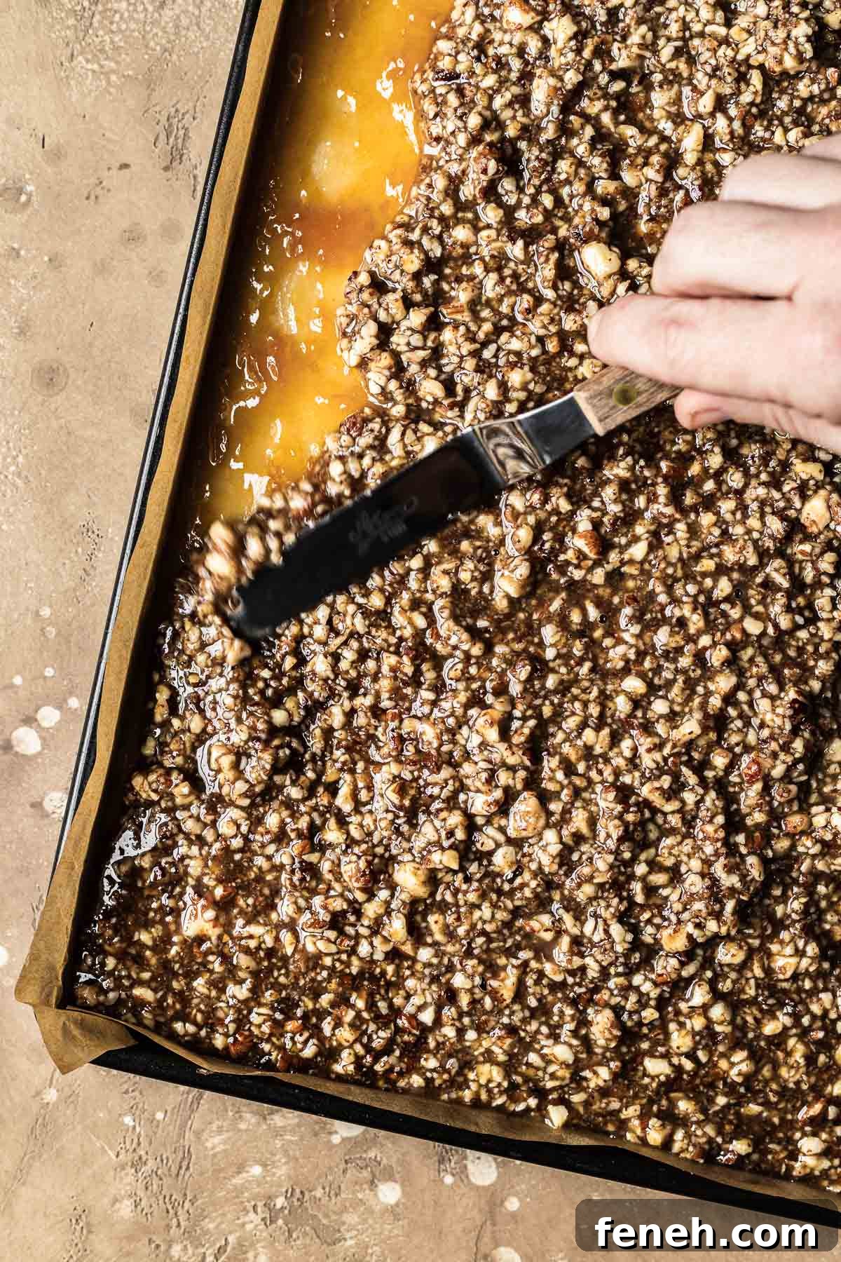 A hand holding a mini spatula spreads nussecken nut topping over apricot jam.