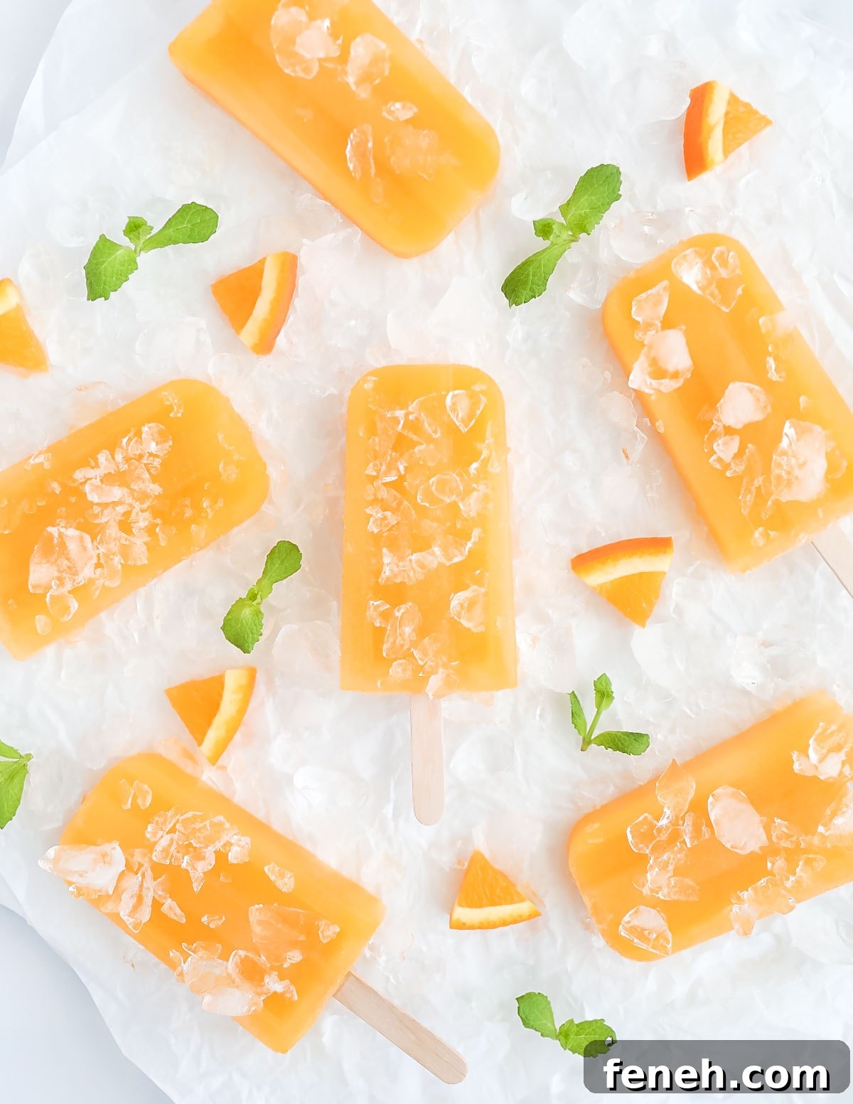 Six orange ice pops on a sheet of white parchment paper, they are covered by and surrounded by crushed pieces of ice, fresh mint leaves, and orange slices