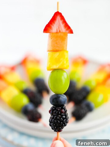 A hand holding up a wooden stick with one piece of strawberry, melon, pineapple, grape, blueberry, and blackberry