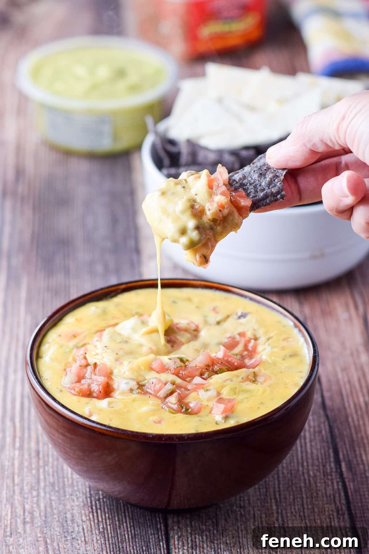 A blue chip in a hand that dipped it in the queso dip with guacamole in the back