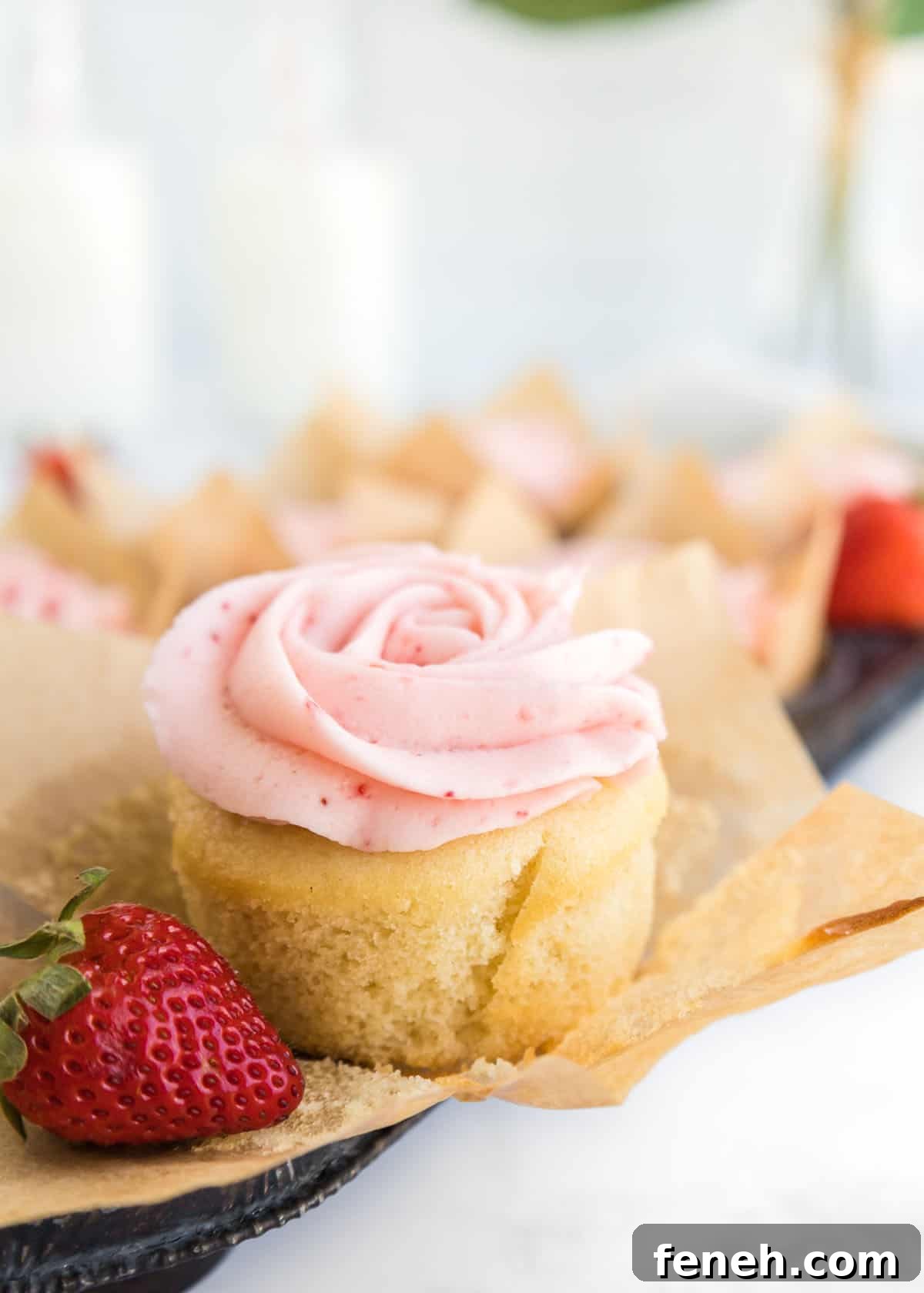 Close-up photo of one cupcake on top of its parchment liner with one whole strawberry in front of it with extra cupcakes in a muffin tin blurred in the background.