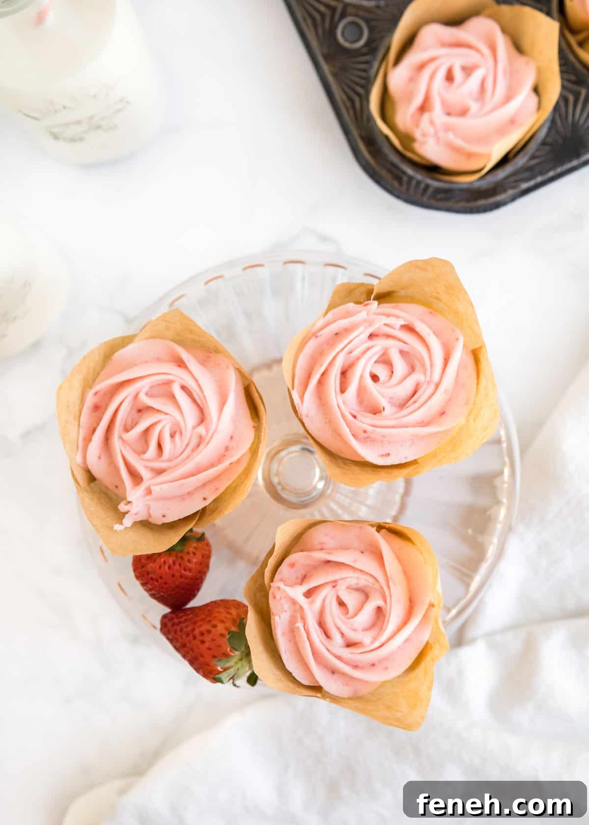 Overhead shot of three strawberry buttercream cupcakes and two whole strawberries on a small, glass cake stand.
